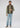 A man stands against a plain background, wearing a green zip-up sweater, the Stelvio Padded Vest in olive green for lightweight warmth, light blue jeans, and gray sneakers.
