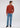 A man is dressed in the Milano 1/4 Zip Merino Wool Sweater in a rust shade, styled with light blue jeans and black shoes. He has short, curly hair, looks down thoughtfully with one hand in his pocket, standing against a simple light background.