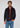 A man stands against a plain light background, looking directly at the camera with a neutral expression. He is wearing a dark jacket over the rust-colored Milano 1/4 Zip Merino Wool Sweater and blue jeans.