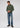 A man stands against a plain background wearing the Milano 1/4 Zip Merino Wool Sweater, layered over a plaid shirt, blue jeans, and brown shoes. He is looking down with his hands clasped in front of him.