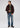 A man stands facing slightly left, wearing a brown jacket, the Milano 1/4 Zip Merino Wool Sweater in green, jeans, and brown shoes. He holds glasses in his right hand and looks to the side against a plain, light-colored background.