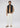 A man stands against a plain background wearing the Milano 1/4 Zip Merino Wool Sweater in black, layered under a black vest and paired with white pants and beige sneakers. He looks straight ahead with one hand in his pocket.