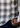 A person’s hand holds the bottom hem of a navy plaid Como Italian Flannel shirt, woven from luxurious Canclini cotton with brown, blue, and white stripes, revealing a small button detail near dark blue jeans.