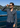 Wearing the Como Italian Flannel—a navy grey plaid shirt crafted from fine Italian flannel—a man in sunglasses stands by a railing overlooking a blue lake, with hills and houses visible in the background beneath a clear sky.
