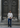A man in a navy plaid Como Italian Flannel shirt and jeans stands on stone steps in front of a large, ornate, dark wooden door with circular carvings.