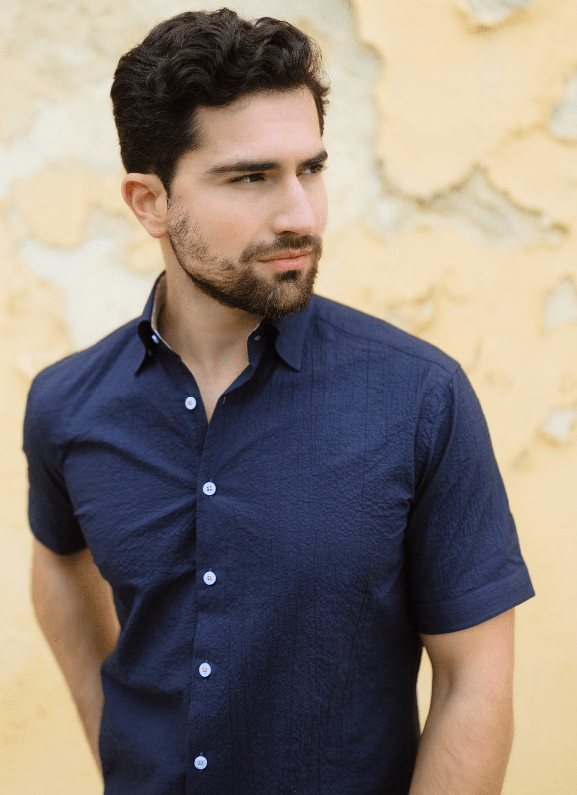 A man with short dark hair and a beard wears the Micah Seersucker Shirt in navy, standing against a textured yellow wall while looking to his left.