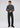 A man stands against a plain background wearing the Milano 1/4 Zip Merino Wool Sweater in dark gray, paired with black jeans and black shoes, looking slightly downward with his hands loosely clasped.