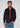 A man with wavy brown hair wears the Stockholm Corduroy Overshirt in navy, layered over a red half-zip sweater and paired with blue jeans. He stands against a plain light background, looking at the camera with a neutral expression—a refined example of versatile layering in men’s fashion.