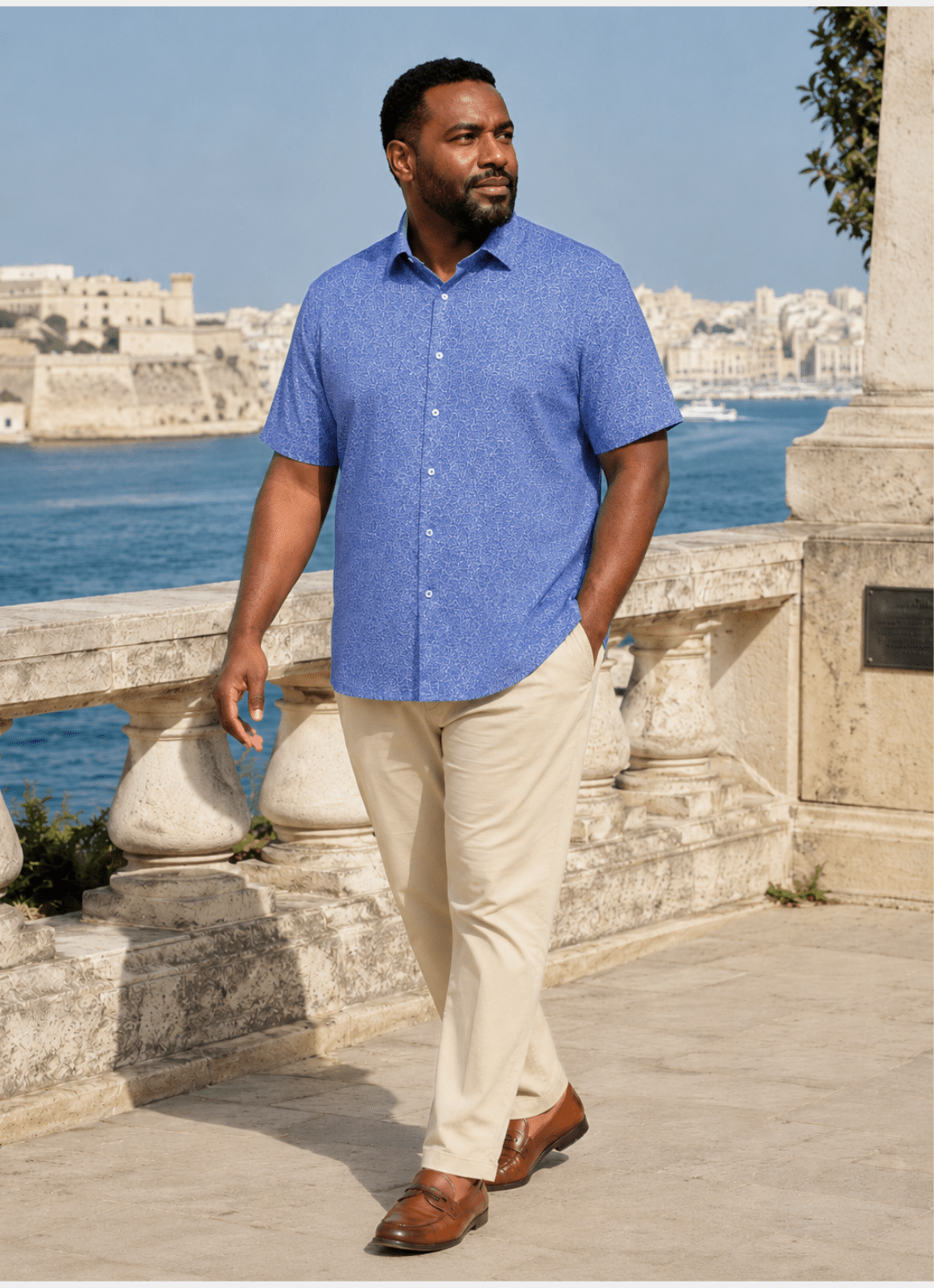 A man wearing the Taunton Stretch Shirt in blue and beige pants walks on a stone terrace by the water, with a cityscape and fortress in the background.